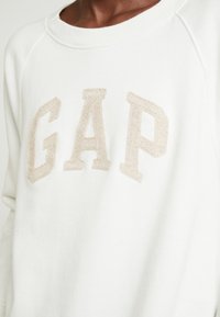 Sudadera blanca con letras "GAP" bordadas en beige, cuello redondo y mangas raglán. El material parece suave y texturizado.