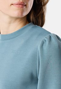 Camicia a maniche lunghe di un azzurro chiaro con colletto rotondo e dettagli delle maniche a sbuffo. "SEASIDE" è discretamente in rilievo sul davanti. Texture morbida.