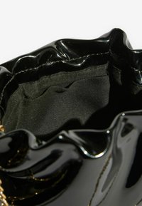 Melna spīdīga soma ar kroku malu un zelta ķēdes detaļu. Interjers izklāts ar gludu melnu audumu, radot mīkstu tekstūru.