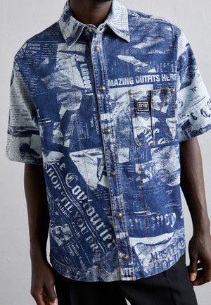 Uomo che indossa una camicia di jeans a maniche corte con bottoni, stampa giornale blu e bianca e una tasca sul petto sul lato sinistro.