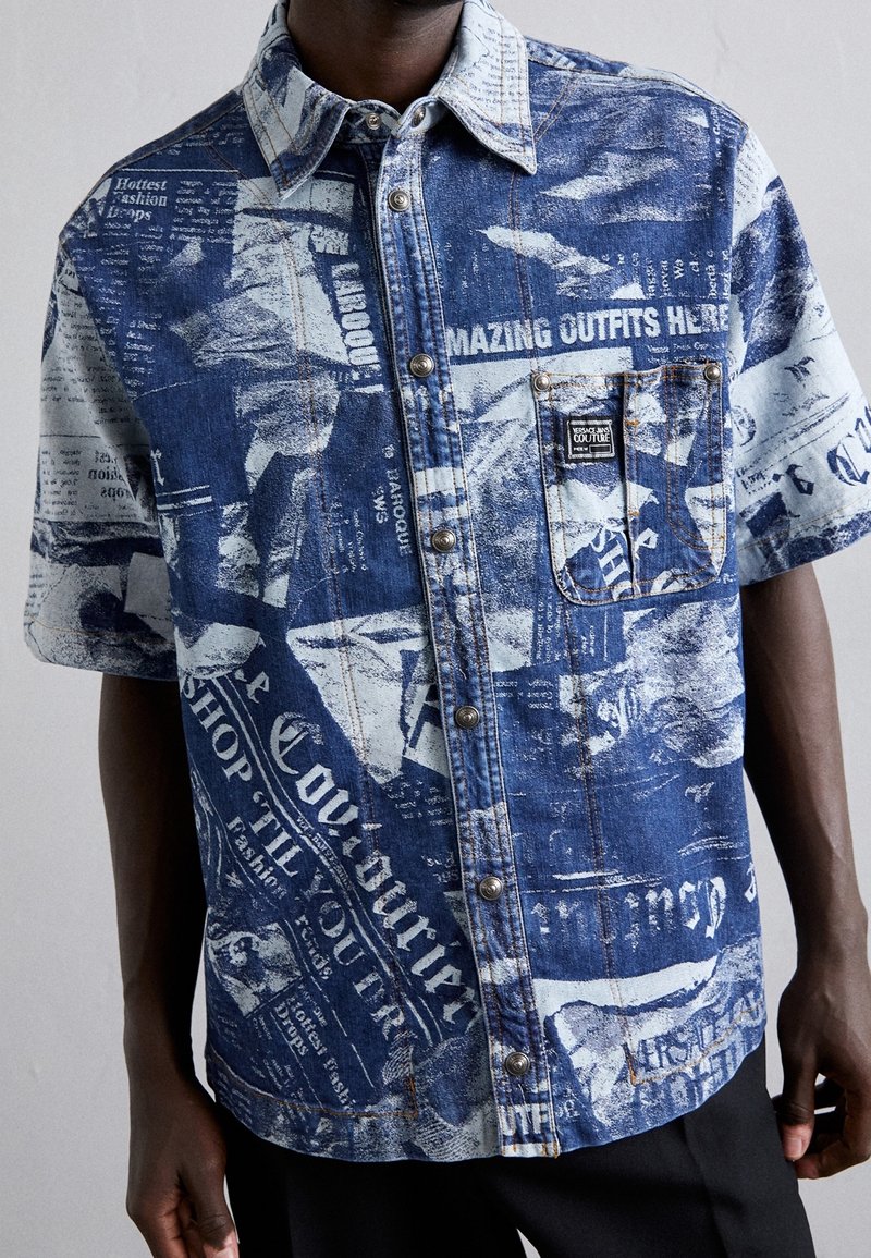 Homme portant une chemise en denim à manches courtes boutonnée avec un imprimé journal bleu et blanc et une poche poitrine sur le côté gauche.