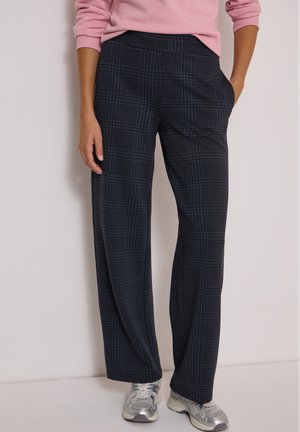 Femme portant un pantalon large à carreaux bleu foncé, un haut à manches longues rose et des baskets argentées, debout avec une main dans la poche.