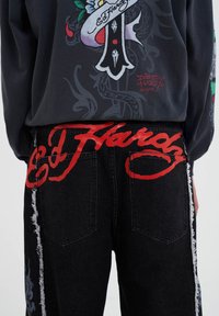 Black denim jeans with red "Ed Hardy" logo on the back, featuring distressed side seams and two rear pockets.