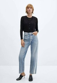 Zwarte button-up vest over een langetermijnsleeve shirt, gecombineerd met lichtblauwe high-waisted jeans met een witte streep, en zwarte gehakte muiltjes.