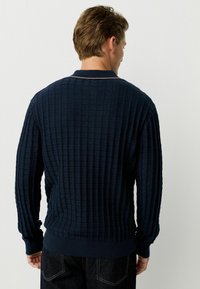 Pulover tricotat texturat de culoare bleumarin, cu un model în grilă, manșete ondulate și un guler cu detalii subtile în dungi.