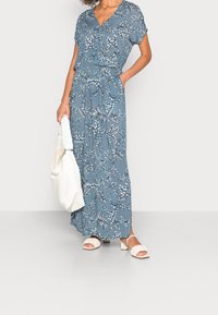 Kvindelig iført en blå blomster-maksikjole med korte ærmer, holder en hvid tote bag og har hvide hæle sandaler på.