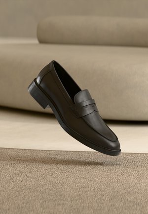 Pantof de tip loafer din piele neagră, odihnindu-se pe un covor bej, cu o canapea curbată bej în fundal.