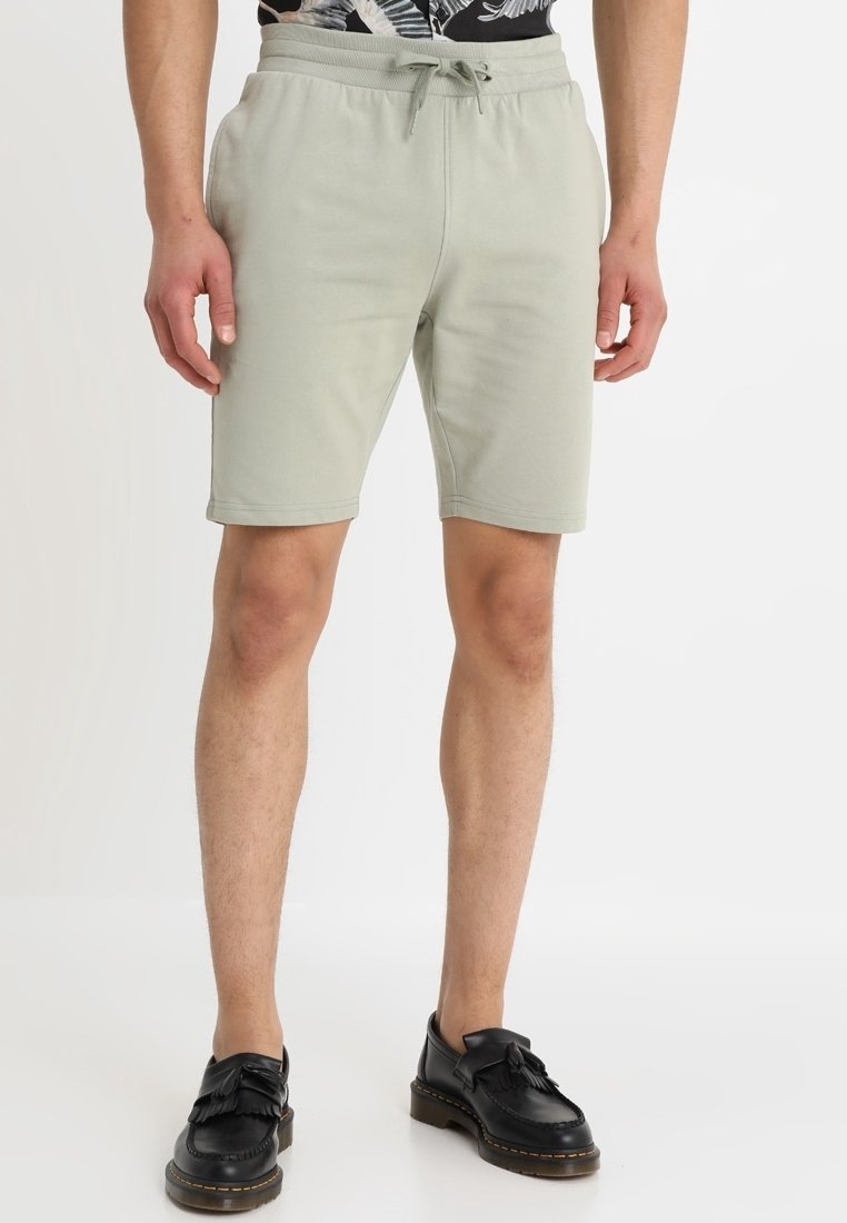Shorts en coton vert clair avec une taille élastique et un cordon de serrage. Longueur genoux, texture lisse, associés à des mocassins à glands noirs.