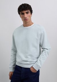 Joven con cabello rizado y oscuro, vistiendo un suéter azul claro sobre una camisa blanca, jeans azul oscuro, posando con una mano en el bolsillo.