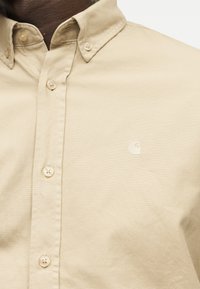 Μπεζ πουκάμισο με κουμπιά, γιακά και κεντημένο λογότυπο στο στήθος, φορεμένο από άτομο με σκουρόχρωμο δέρμα.