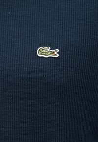 Μικρό πράσινο λογότυπο κροκοδείλου με ανοιχτό κόκκινο στόμα κεντημένο σε σκούρο μπλε υφή υφάσματος.