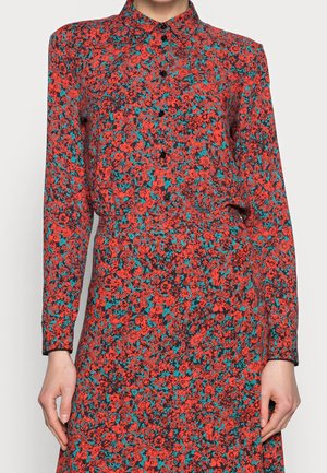 Kobieta ubrana w sukienkę na guziki z długim rękawem, ozdobioną gęstym wzorem kwiatowym w kolorze czerwonym i błękitno-zielonym na czarnym tle.