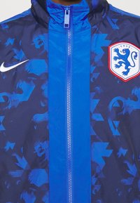 Nike vindjacka i blå nyanser med ett geometriskt mönster, dragkedja och KNVB-emblem med lejonlogotyp på bröstet.