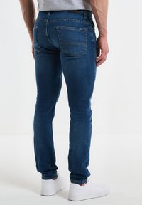 Kitsad sinised teksad sujuva tekstuuri ja minimaalse kulumisega, millel on neli taskut ja kollane niit, koos valgete tossudega.