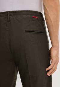 Ciemnozielone spodnie mają elastyczny pas, tylną kieszeń z wąskim otworem oraz czerwoną metkę marki blisko szwu pasa.