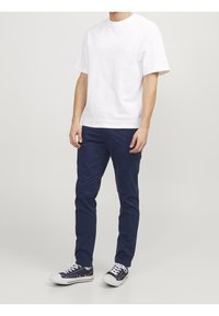 Homme portant un t-shirt blanc uni à manches courtes, un pantalon slim-fit bleu marine et des baskets noir et blanc, se tenant contre un fond clair.