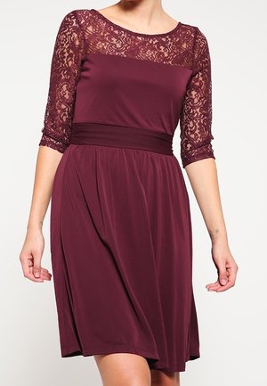 Kobieta w bordowej sukience do kolan z koronkową górą i rękawami do połowy przedramienia, z talią podkreśloną paskiem, stojąca na tle jednolitego tła.