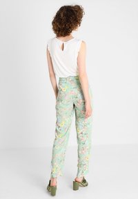 Bílý bezrukávový top s volánovými rukávy a průstřihem na zádech, spárovaný s kalhotami s květinovým vzorem v světle zelené barvě a zelenými sandály na vysokém podpatku.