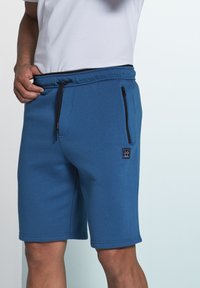 Blaue Sweatshorts aus weichem Stoff mit einem elastischen Bund, Kordelzug und einer seitlichen Reißverschlusstasche. Mit einem kleinen Logo-Patch.