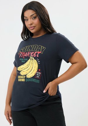 Kvinde iført sort "Sunday Market" T-shirt med banangrafik og tekst om frisk, lokalt dyrkede produkter.