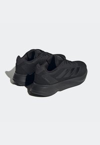 Une paire de chaussures de sport noires avec une texture en maille, des semelles épaisses et un design à lacets, montrées sous un angle latéral arrière sur un fond clair.