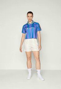 Camiseta deportiva recortada en azul y púrpura con cuello amarillo, logo blanco y pantalones cortos blancos. Llevado con zapatillas y calcetines blancos.