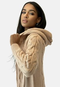 Mjuk kabelstickad hoodie i beige med ett strukturerat mönster, förlängda ärmar och huva med dragsko, som erbjuder en avslappnad passform.