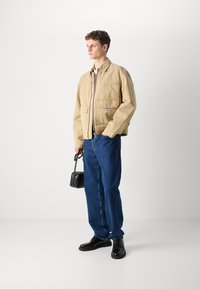 Beige bomulljakke med frontlommer, sammen med blå denimjeans og svarte lærsko. Holder en liten svart håndveske.