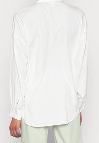 Bílá košile s dlouhým rukávem a hladkou texturou, s knoflíkovými manžetami a zakřiveným lemem. Pohled zezadu ukazuje minimalistický design bez viditelných vzorů.