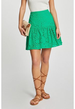 Kvinde iført grøn nederdel med hulmønster, hvid tekstureret top, holder en beige clutch og brune snøresandaler med flad sål.