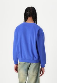 Sudadera azul de corte relajado, con puños y dobladillo acanalados, textura suave y un clásico escote tipo crew; combinada con jeans de lavado claro.