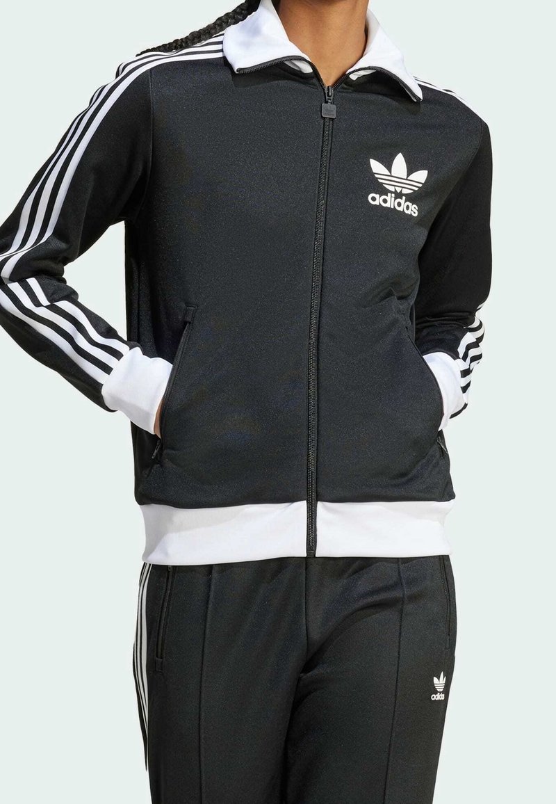 Persoon in een zwart Adidas trainingspak met witte strepen op de mouwen en broek, handen in de zakken van de jas, staand tegen een lichte achtergrond.