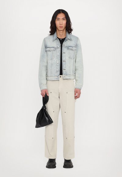 Light denim jacket, black top, beige wide-leg pants, black footwear, holding a black fabric bag. Jacket features metal buttons and chest pockets.