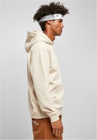 Krém színű kapucnis pulóver elülső zsebbel, puha tapintással és bordázott mandzsettával, barna nadrággal párosítva, amelyen oldalt márkajelzés található.