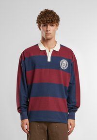 Rugby polo s dlhým rukávom v maroon a námorníckych pruhoch, s bielym golierom a vyšitým logom na hrudi. Vyrobené z mäkkého materiálu.