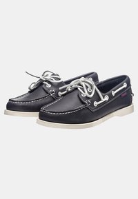Zapatos náuticos de cuero negro con costuras y cordones blancos, que cuentan con una suela de goma blanca y ojales plateados para la sujeción de los cordones.