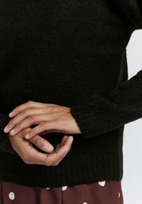 Schwarzer gestrickter Pullover aus strukturiertem Stoff. Mit langen Ärmeln und einem gerippten Saum. Die Hände sind sanft vor dem Kleidungsstück gefaltet.