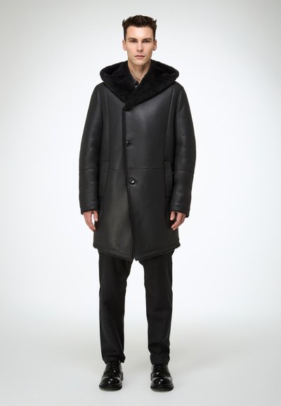 Schwarzer Ledermantel mit großer, strukturierter schwarzer Pelzkapuze. Verfügt über Knopfverschlüsse und Seitentaschen. Wird über schwarze Hosen und Schuhe getragen.