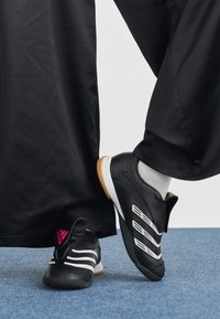 Person som har på seg svarte Adidas innefotballsko med hvite striper og gummisåler, stående på blått teppe med svarte vide bukser.