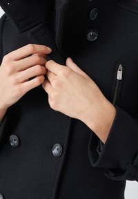 Cappotto nero doppiopetto in tessuto strutturato. Presenta grandi bottoni in plastica, un colletto alto e una tasca con zip sul lato.