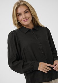 Blonde Frau lächelt, trägt eine lockere schwarze Bluse mit Knöpfen und schmalen Puffärmeln sowie kleine Creolen, vor einem schlichten, hellen Hintergrund.