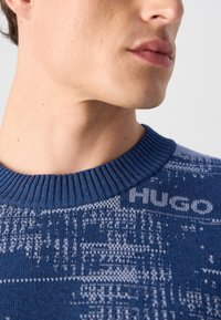 Sininen neulepusero, jossa on ribbireunus ja tekstuurinen kuvio, sekä vaaleanharmaa "Hugo" -logo olkapään alueella.