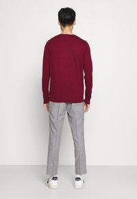 Burgunderfarbene Strickpullover mit langen Ärmeln, kombiniert mit hellgrauen, maßgeschneiderten Hosen. Das Model steht vor einem schlichten Hintergrund und trägt weiße Sneaker.