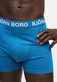 Κοντινό πλάνο ενός ατόμου που φοράει μπλε boxer Björn Borg με το όνομα της μάρκας ορατό στη μέση, το χέρι ακουμπάει στο ισχίο.