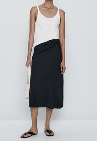 Donna che indossa un top bianco senza maniche e una gonna midi nera con arricciature laterali, sandali neri e tiene una borsa beige al suo fianco.