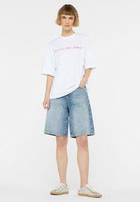 White oversized t-shirt with pink lettering paired with light blue denim shorts. Model wears silver sneakers with beige accents. Simple, casual style.