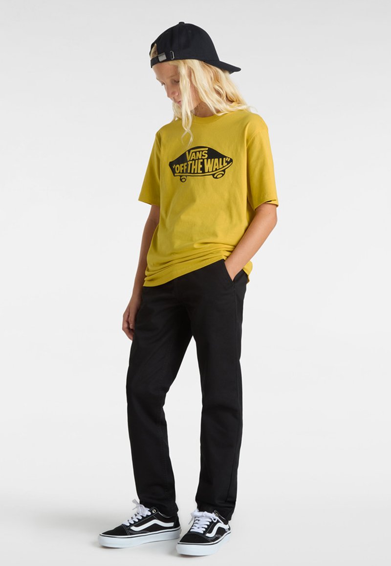 Gul kortärmad t-shirt med svart "VANS OFF THE WALL" logotyp, tillsammans med svarta byxor och svart-vita sneakers. Svart keps bärs bakåtvänd.