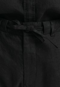 Pantalon en lin noir avec une ceinture en tissu nouée par un simple nœud à la taille, montrant des passants de ceinture et des détails de couture.