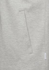 Tissu gris clair présentant une texture mate avec une poche horizontale épurée et une petite étiquette de marque sur le côté.