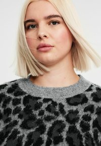 Harmaa neule, jossa on mustia leopardi-pilkkuja, pyöreä pääntie ja tekstuurinen kangas. Mallilla on vaaleat hiukset ja neutraali tausta.